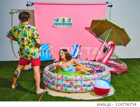 Young man in colorful Hawaiian shirt playfully sprays water at woman in retro swimsuit lounging in inflatable pool. 114736754