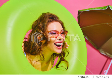 Portrait of young woman holds neon-green inflatable ring and smiles brightly peeking out of it against bright pink background. Portrait of young woman holds neon-green inflatable ring and smiles brightly peeking out of it against bright pink background. 114736785