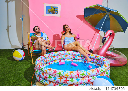 Man and woman enjoying their backyard beach setup, sitting by inflatable pool, toasting with drinks. Summer mood. Man and woman enjoying their backyard beach setup, sitting by inflatable pool, toasting with drinks. Summer mood. 114736793