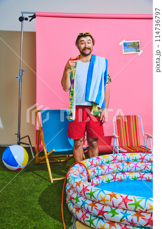 Man dressed vibrant shorts and shirt holding refreshing cocktail standing near inflatable pool against studio background. 114736797