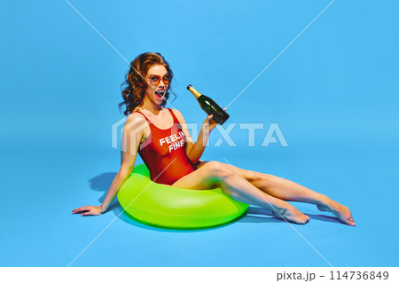 Happy young woman dressed in retro swimsuit drinks champagne resting in neon colored inflatable ring against blue background. 114736849