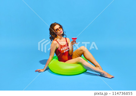 Beautiful, slim woman in retro swimwear sitting in neon colored inflatable ring with cocktail against blue background. Beautiful, slim woman in retro swimwear sitting in neon colored inflatable ring with cocktail against blue background. 114736855