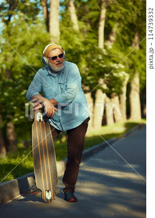Senior Style. Elderly man in stylish clothes, beanie hat, headphones and sunglasses standing in greenery park with skateboard. Senior Style. Elderly man in stylish clothes, beanie hat, headphones and sunglasses standing in greenery park with skateboard. 114739332