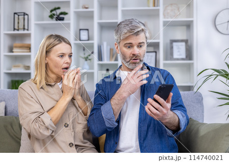 Middle-aged couple with shocked expressions, looking at a smartphone while sitting on the couch in a modern living room. 114740021