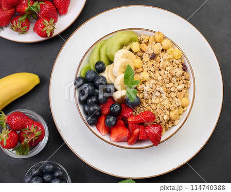 Granola with strawberries, kiwi, banana and blueberries in a round plate on a black table. Healthy and tasty food Granola with strawberries, kiwi, banana and blueberries in a round plate on a black table. Healthy and tasty food 114740388