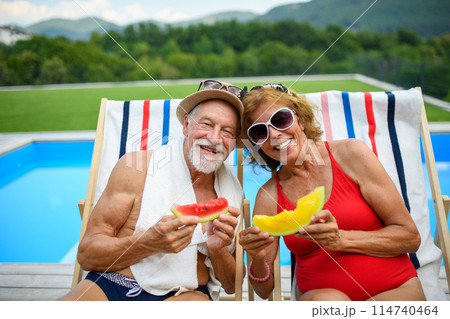 Beautiful elderly couple enjoying their vacation. Seniors having romantic time, sitting by swimming pool in lounche chairs sunbathing. 114740464