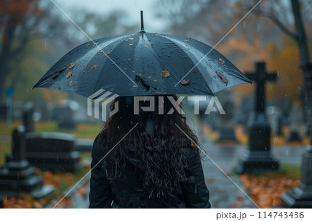Selective focus of Back view of woman wearing black dress holding umbrella on rainy day at Christian cemetery. 114743436