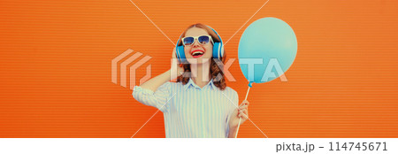 happy cheerful young woman in headphones listening to music with blue balloon on orange background 114745671
