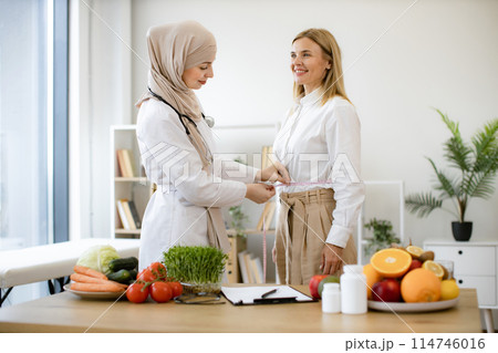 Muslim woman in doctor's coat and hijab finding out waist length of female. 114746016