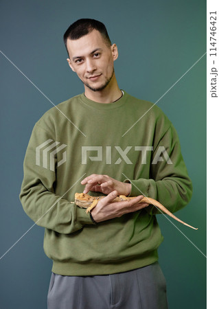 Vertical portrait of Asian young man holding iguana in hands carefully standing against green background and looking at camera 114746421