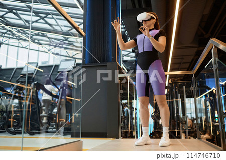 Full length portrait of sportive young woman wearing VR headset in gym and using interactive technology with hand tracking copy space 114746710