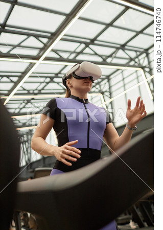 Vertical low angle portrait of sportive woman running on treadmill and wearing VR headset in futuristic gym 114746740