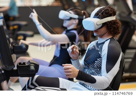 Side view portrait of young man on exercise bikes in gym wearing VR headset for immersive training simulation copy space Side view portrait of young man on exercise bikes in gym wearing VR headset for immersive training simulation copy space 114746769