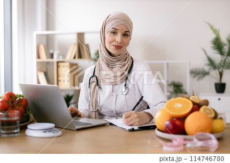 Muslim female nutritionist sitting and working with laptop in modern clinic. Muslim female nutritionist sitting and working with laptop in modern clinic. 114746780