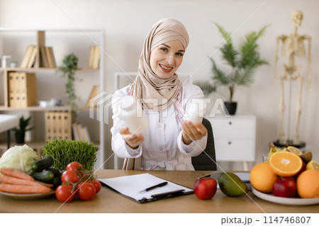 Portrait of positive Muslim lady in hijab and lab coat smiling at camera. Portrait of positive Muslim lady in hijab and lab coat smiling at camera. 114746807