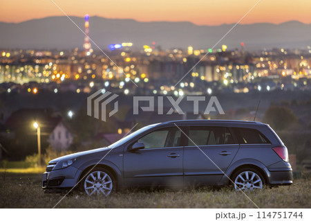 Gray car parked at night in green meadows on background of lights of distant city buildings and dark mountain ridge under bright sky at sunset 114751744