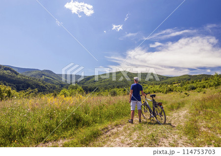 Young man standing near bicycle in morning sunrise with wonderful rays and morning mist during calm summer active day Young man standing near bicycle in morning sunrise with wonderful rays and morning mist during calm summer active day 114753703