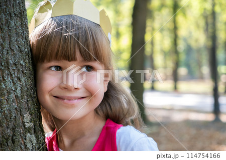 Portrait of a pretty smiling child girl in a paper crown on her head leaning to a tree in summer park. Portrait of a pretty smiling child girl in a paper crown on her head leaning to a tree in summer park. 114754166