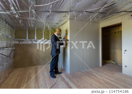 Strange man in businessman suit and gas protection mask inside a room under renovation works holding electric screwdriver and a level tools. 114754186