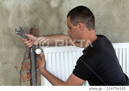 Young handsome professional plumber worker installing heating radiator on brick wall using a wrench in an empty room of a newly built apartment or house. Construction, maintenance and repair concept. Young handsome professional plumber worker installing heating radiator on brick wall using a wrench in an empty room of a newly built apartment or house. Construction, maintenance and repair concept. 114754195