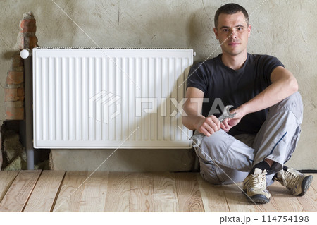 Young handsome plumber sitting on floor with wrench in hand near successfully installed heating radiator in empty room of a newly built apartment or house. Construction, maintenance and repair concept Young handsome plumber sitting on floor with wrench in hand near successfully installed heating radiator in empty room of a newly built apartment or house. Construction, maintenance and repair concept 114754198