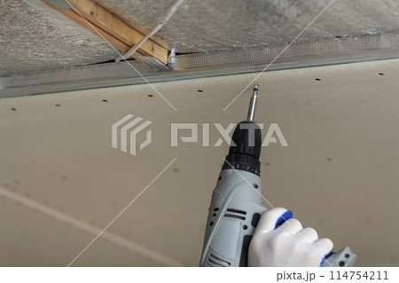 Close-up of constructor worker hand in protective glove with electrical cordless screwdriver connecting suspended drywall ceiling to metal frame. Renovation, construction and DIY concept. Close-up of constructor worker hand in protective glove with electrical cordless screwdriver connecting suspended drywall ceiling to metal frame. Renovation, construction and DIY concept. 114754211