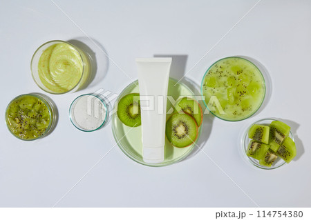 Flat lay glassware containing fresh kiwi, green essence and yellow exfoliating scrubs above, green color of kiwi striking on white countertop. Unlabeled cosmetic tube in center, space for text Flat lay glassware containing fresh kiwi, green essence and yellow exfoliating scrubs above, green color of kiwi striking on white countertop. Unlabeled cosmetic tube in center, space for text 114754380