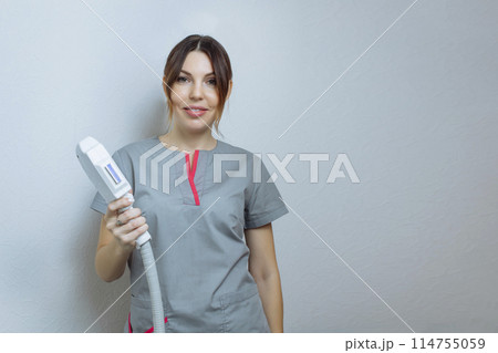 Female cosmetologist in a medical coat holds a working part of the modern laser epilator in her hands and poses for a photo. Laser epilation treatment in cosmetic beauty clinic. 114755059