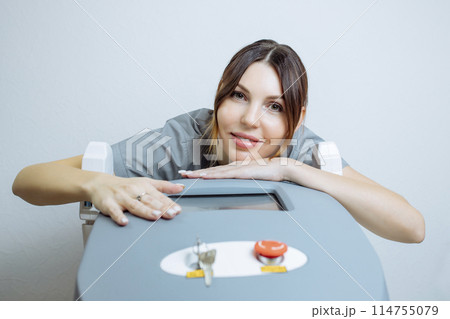 Female cosmetologist in a medical coat holds a working part of the modern laser epilator in her hands and poses for a photo. Laser epilation treatment in cosmetic beauty clinic. 114755079