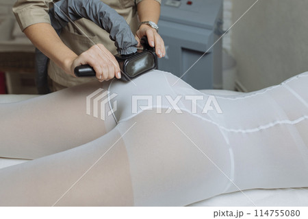 Close up shot of therapist beautician makes LPG massage treatment to young woman in white transparent suit, lying at beauty SPA clinic. Focus on hands holding medical unit 114755080