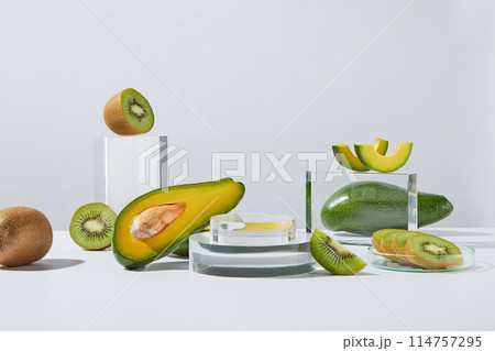 Photo was taken from front view over white background, an empty glass pedestal in center, decorated with fresh kiwis, avocados and rectangular transparent podium. Space for advertising 114757295