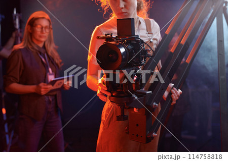 Cropped shot of young woman operating video camera on rig in studio with neon lights copy space Cropped shot of young woman operating video camera on rig in studio with neon lights copy space 114758818