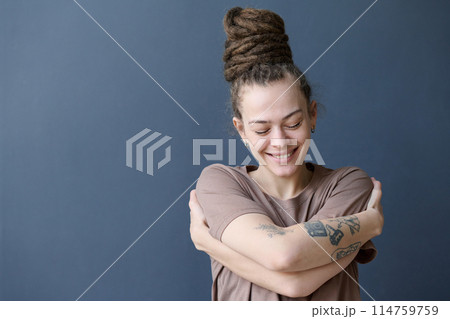 Candid portrait of smiling Caucasian woman with dreadlocks in bun hairstyle embracing herself standing by grey background copy space Candid portrait of smiling Caucasian woman with dreadlocks in bun hairstyle embracing herself standing by grey background copy space 114759759