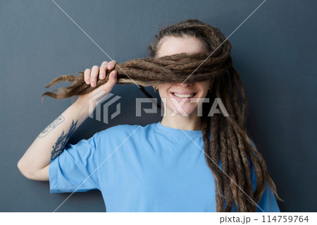 Portrait of tattooed Caucasian woman with long dreadlocks style smiling standing by wall hair covering eyes 114759764