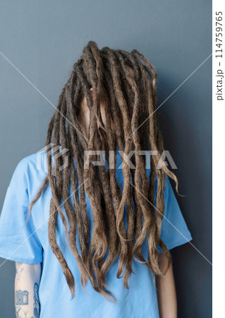 Vertical portrait of tattooed Caucasian woman with long dreadlocks style standing by wall hair covering face 114759765