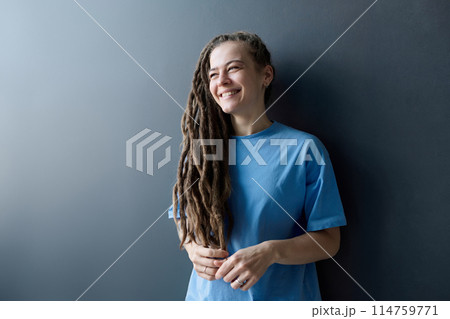 Minimal waist up portrait of young Caucasian woman with long dreadlocks standing by grey wall and looking away with smile copy space 114759771