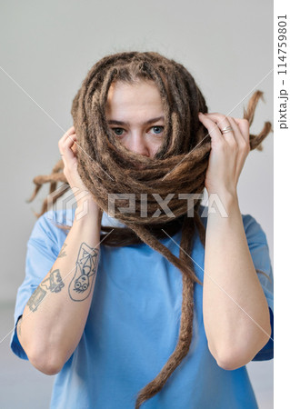 Minimal portrait of young Caucasian woman with long dreadlocks hairstyle wrapping face with hair and looking at camera 114759801