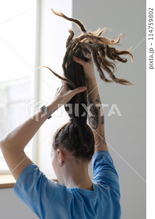 Vertical back view of young Caucasian woman holding long dreadlocks in ponytail facing window 114759802