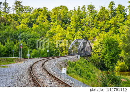 A curved railway bridge in Rataje surrounded by dense greenery and trees during summer, with railway tracks leading up to the bridge. 114761159