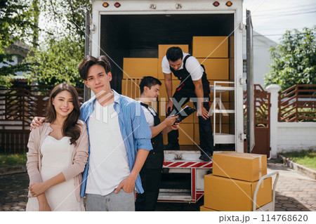 Captured in a portrait a smiling couple watches professional movers load furniture onto a truck for relocation. Quality service and teamwork for happy customers. Moving Day Concept Captured in a portrait a smiling couple watches professional movers load furniture onto a truck for relocation. Quality service and teamwork for happy customers. Moving Day Concept 114768620