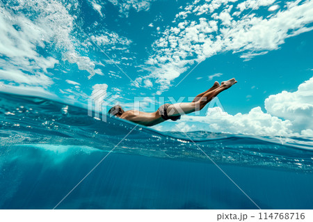 An athletic man dives gracefully into the clear blue ocean beneath a bright, cloud-dappled sky 114768716