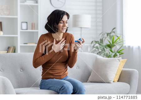 Middle-aged woman sits on sofa, looking distressed as she experiences chest pain and holds an asthma inhaler, suggesting a breathing difficulty situation in a living room setting. Middle-aged woman sits on sofa, looking distressed as she experiences chest pain and holds an asthma inhaler, suggesting a breathing difficulty situation in a living room setting. 114771885