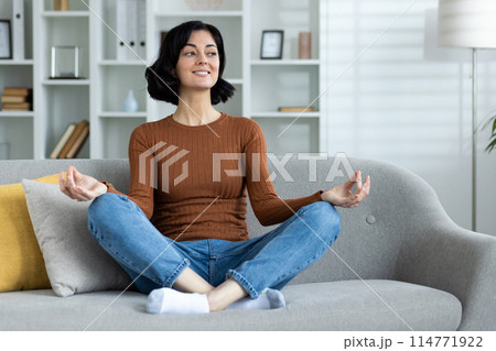 Young woman sitting on a sofa in a living room practicing meditation. Relaxed and peaceful ambiance. Young woman sitting on a sofa in a living room practicing meditation. Relaxed and peaceful ambiance. 114771922