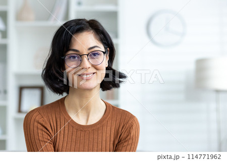 Portrait of a smiling woman wearing glasses and an orange sweater, sitting in a modern home office setting with white shelves and a clock in the background. Portrait of a smiling woman wearing glasses and an orange sweater, sitting in a modern home office setting with white shelves and a clock in the background. 114771962