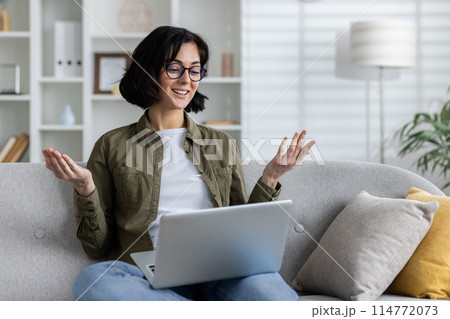A young smiling woman in casual clothes is sitting on the sofa at home, holding a laptop on her lap and talking on a video call while gesturing with her hands. A young smiling woman in casual clothes is sitting on the sofa at home, holding a laptop on her lap and talking on a video call while gesturing with her hands. 114772073