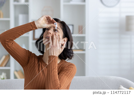 Close-up photo of a young woman sitting on the sofa at home and instilling medicine for fatigue and inflammation in her eyes. Close-up photo of a young woman sitting on the sofa at home and instilling medicine for fatigue and inflammation in her eyes. 114772117