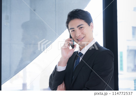 窓際で笑顔で電話(通話)をするスーツを着た男性ビジネスマン　カメラ目線 114772537