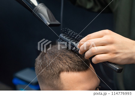 Hairdresser dries hair with a hairdryer and comb to a client in a barbershop. Hairdresser dries hair with a hairdryer and comb to a client in a barbershop. 114776993