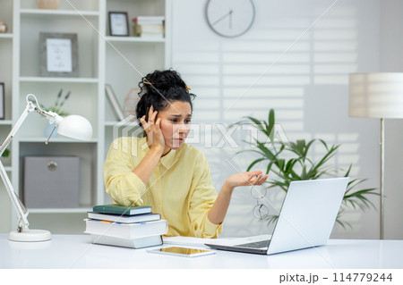 Woman feeling frustrated while working on a laptop in a home office setting with books and paperwork. 114779244