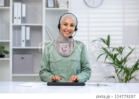 Smiling customer service representative wearing a headset and hijab while working in a modern office environment, ready to assist customers. 114779287
